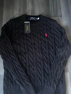 Svart kabelstickad tröja Polo Ralph Lauren - Kabelstickad svart tröja från Polo Ralph Lauren med rund halsringning och den klassiska röda logotypen broderad på bröstet. Tröjan har ribbade muddar vid ärmslut och nederkant. Perfekt för dig som gillar stilrena och tidlösa plagg.