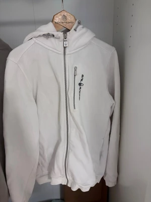 Vit hoodie från Sail Racing - Säljer en vit hoodie från Sail Racing med dragkedja framtill och en bröstficka med logga. Hoodien har justerbar huva med snören och en liten broderad logga på huvan. Tillverkad i mjukt material, perfekt för en avslappnad stil. Den är mycket bra skick förutom att luvan är lite missfärgad på insidan, kan försvinna med rätt tvättmedel ej provat, inget som syns när man har på sig den