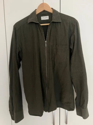Olivgrön overshirt från John Henric - Stilren olivgrön overshirt från John Henric med dragkedja framtill, klassisk krage och bröstficka. Tillverkad i ett mjukt tyg som ger en avslappnad look. Perfekt att bära över en t-shirt eller skjorta för en trendig lager-på-lager stil.