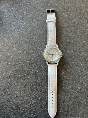 Vit klocka med strass och läderband - Stilren klocka från Geneva med vitt läderband i krokopräglad design och silverfärgat ur med strass runt boetten. Klassisk urtavla med tydliga siffror och detaljer i silver. Perfekt accessoar för att lyfta din outfit. Behöver nytt batteri