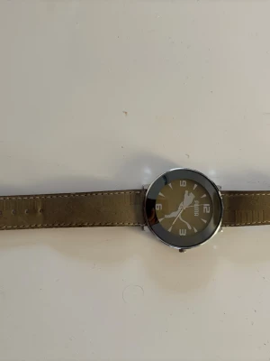 Beige klocka från Puma med läderband - Stilren klocka från Puma med beige läderband och rund silverfärgad boett. Urtavlan är beige med vita siffror och Puma-loggan synlig. Perfekt accessoar för dig som gillar sportig och enkel design.