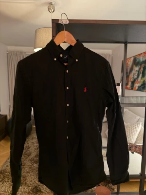 Svart skjorta från Ralph Lauren - Klassisk svart skjorta från Ralph Lauren med knappkrage och den ikoniska röda logotypen broderad på bröstet. Skjortan har långa ärmar och är tillverkad i 100% bomull, vilket gör den både bekväm och stilren. Perfekt för dig som gillar tidlös design.
