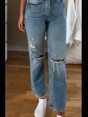 Ljusblå raka jeans med slitningar - Säljer ett par ljusblå jeans med raka ben och rejäla slitningar över knäna och låren. Jeansen har hög midja och klassisk femficksmodell. Perfekta för en avslappnad och trendig look.