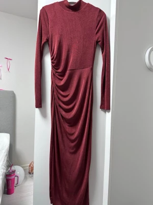 Vinröd långklänning med rynk - Snygg vinröd långklänning med långa ärmar och hög hals. Klänningen har en draperad detalj på sidan som ger en cool rynkeffekt. Tillverkad i ett mjukt, stretchigt material som sitter bekvämt på kroppen. Perfekt för dig som vill sticka ut med stilren elegans.