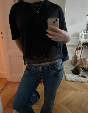 Svart croppad topp med spetskant - Svart croppad topp med korta ärmar och dekorativ spetskant nertill. T-shirten har en loose passform och är perfekt för en avslappnad stil. Materialet känns mjukt och lätt, vilket gör den bekväm att bära.