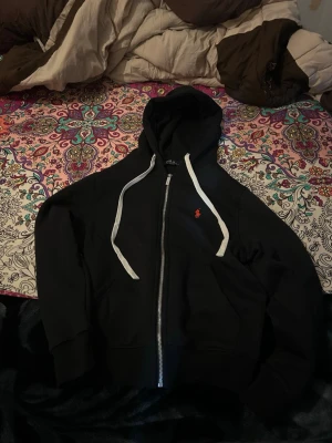 Svart zip hoodie från Polo Ralph Lauren. - Svart hoodie med dragkedja från Polo Ralph Lauren. Klassisk design med vit dragsko i huvan och röd broderad logga på bröstet. Mjuk bomullskänsla och kängurufickor framtill. Perfekt för en chill och stilren look.