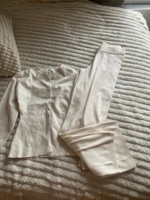 Krämvitt loungewear-set med knappar - Mysigt loungewear-set i krämvitt med långärmad tröja och matchande byxor. Tröjan har rund halsringning och knappar framtill, byxorna har ribbade muddar. Perfekt för chill dagar hemma. Mjukt stickat material för extra komfort.
