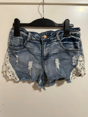 Blå jeansshorts med spetsdetaljer strl: 36 - Snygga blå jeansshorts från Rut & Cirkle med slitna detaljer och vit spets längs sidorna. Shortsen har råa kanter, klassisk femficksdesign och knappstängning. Perfekta för varma dagar och en avslappnad stil.