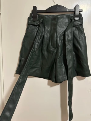 Mörkgröna skinnshorts med knytband strl: 32 - Snygga mörkgröna shorts i skinnimitation från amisu Modellen är högmidjad med breda ben och har ett långt knytband i midjan för extra stil. Perfekta för dig som vill ha en edgy look med trendiga detaljer.