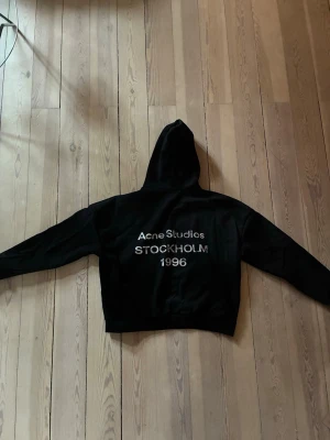 Svart hoodie Acne Studios Stockholm - Svart hoodie från Acne Studios med stor huva och tryck i vitt på ryggen där det står 'Acne Studios STOCKHOLM 1996'. Klassisk passform med ribbade muddar. Perfekt för dig som gillar streetwear och vill ha något stilrent men ändå statement.