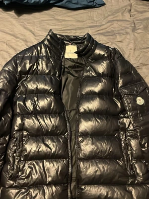 Svart dunjacka från Moncler - Tja! Säljer nu min äldre svarta dunjacka från Moncler med glansig finish och quiltad design. Jackan har ståkrage, dragkedja framtill och två sidofickor med dragkedja. Moncler loggan på ärmen och innerfoder med serietidningsmönster. Jackan är som sagt gammal därav priset men är fortfarande behållen i ett väldigt gott skick! Vid frågor så är det bara att ställa!