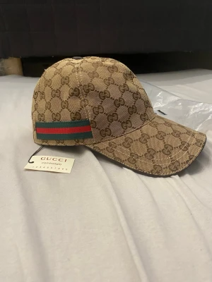 Gucci keps med GG-mönster - Snygg beige keps från Gucci med klassiskt GG-monogram i brunt över hela kepsen. På sidan finns den ikoniska gröna och röda randen. Kepsen har böjd skärm och justerbar rem bak. Perfekt för dig som vill ha en lyxig streetstyle-look. Du får boxen med.