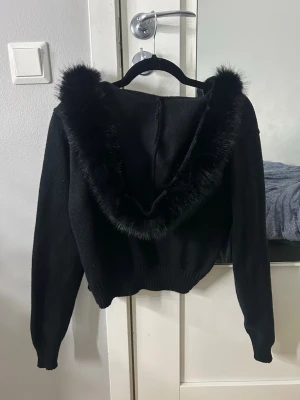 Svart stickad hoodie med fuskpäls - Snygg svart stickad hoodie med luva kantad av mjuk svart fuskpäls. Tröjan har långa ärmar och ribbade muddar vid ärmslut och midja. Perfekt för dig som vill ha en trendig och mysig look.