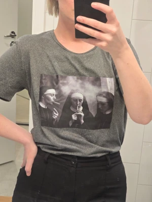 Grå t-shirt med tryck av nunnor - Gråmelerad t-shirt med ett svartvitt tryck på bröstet som föreställer tre nunnor som röker. T-shirten har rund halsringning och korta ärmar. Perfekt för dig som gillar unika och lite edgy motiv. Står M men tycker den passar S bättre