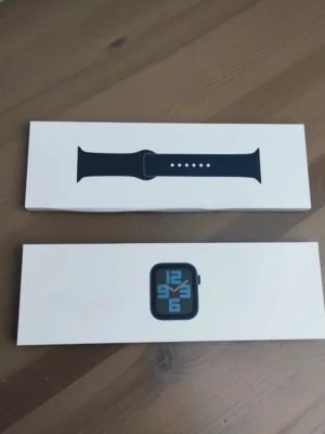 Apple Watch SE 38mm - Säljer en Apple Watch SE 38mm i svart aluminium med matchande svart sportarmband. Klockan har en stilren, fyrkantig design och kommer i originalförpackning. Perfekt för dig som vill ha koll på notiser, träning och hälsa direkt på handleden.