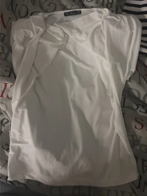 Vit topp med knyt från Tille - Säljer en vit topp från Tille med korta ärmar och snygg knyt-detalj vid axeln. Toppen har en enkel och clean look, perfekt till jeans eller kjol. Materialet känns mjukt och lätt, troligen bomull eller polyester.