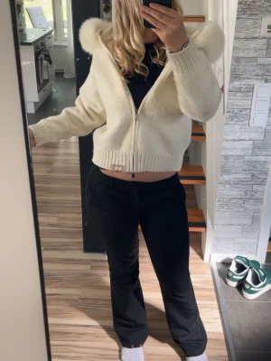 Vit stickad hoodie med fuskpäls - Säljer en vit stickad hoodie med dragkedja och fluffiga fuskpälsdetaljer på huvan. Tröjan har ribbstickade muddar och är tillverkad i mjukt material som ger en cozy känsla. Perfekt för dig som vill ha en trendig och varm look.