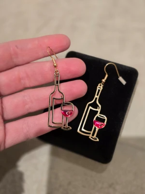Guldörhängen med vinflaska och glas - Unika örhängen i guldfärgad metall formade som vinflaskor och vinglas med röd emaljdetalj i glaset. Perfekta för dig som vill ha något lekfullt och trendigt. Krokar i metall för enkel användning. Säljs som par.