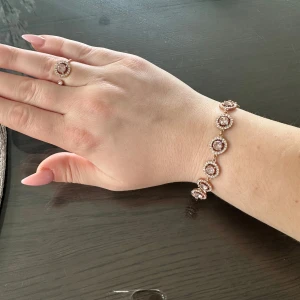Glittrigt armband och ring i roséguld - Säljer ett matchande set med armband och ring i roséguld. Båda har runda, ljusrosa stenar i mitten omgivna av små gnistrande stenar för extra bling. Perfekt för dig som gillar eleganta och glittriga accessoarer med lyxig känsla.