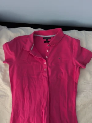 Rosa pikétopp från Tommy Hilfiger - Säljer en snygg rosa pikétopp från Tommy Hilfiger i slim fit-modell. Toppen har korta ärmar, klassisk krage och knappar framtill. Liten logga på bröstet och mjukt bomullsmaterial som känns skönt mot huden.