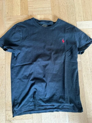 Svart t-shirt från Polo Ralph Lauren - Klassisk svart t-shirt från Polo Ralph Lauren med röd broderad logga på bröstet. T-shirten har rund halsringning och korta ärmar. Tillverkad i mjuk bomull för en skön känsla och enkel stil. 