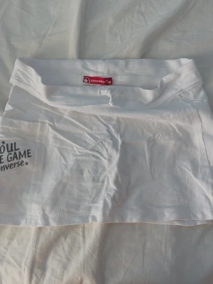 Vit skort från Converse - Snygg vit skort från Converse med trycket 'SOUL THE GAME Converse' på sidan. Skorten har en sportig look med bred linning och är gjord i ett mjukt, stretchigt material som känns bekvämt. Perfekt för dig som gillar streetwear och vill ha något unikt.