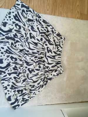 Kort kjol med zebramönster - Säljer en kort kjol med svartvitt zebramönster och volangdetaljer. Kjolen har resår i midjan för skön passform och är gjord i ett lätt, syntetiskt material. Perfekt för dig som vill sticka ut med en trendig look.