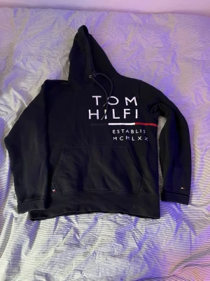 Svart hoodie från Tommy Hilfiger - Svart hoodie från Tommy Hilfiger med stor vit logga och text på både fram- och baksidan. Klassisk känguruficka framtill, huva med dragsko och diskreta röda och vita detaljer. Tillverkad i mjukt bomullsmaterial för en skön och avslappnad look.