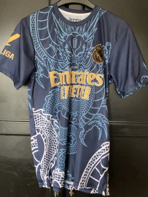 Real Madrid Ronaldo matchtröja blå - Snygg blå matchtröja från Real Madrid med stort drakmotiv i ljusblått och vita detaljer. Tröjan har gula tryck med Emirates Fly Better på framsidan och Ronaldo 7 på ryggen. Korta ärmar och rund hals. Perfekt för fotbollsfans!