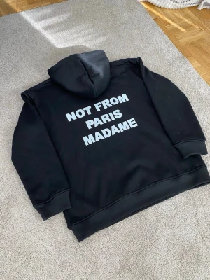 Drole De Monsieur Not From Paris Madame Hoodie - Svart hoodie från Drole De Monsieur Not From Paris Madame med stor vit text på ryggen. Hoodien har huva och ribbade muddar vid ärmslut och nederkant. Insidan är mjuk och borstad för extra komfort. Perfekt för dig som gillar streetwear och statement-plagg. Storlek M men mindre i storlek passar mer som S.