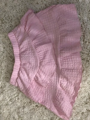 Rosa volangkjol med struktur - Supersöt rosa kortkjol med volanger och strukturerat tyg. Kjolen har bred resår i midjan och dubbla volanglager som ger en lekfull look. Perfekt för dig som vill ha en feminin och trendig stil💕