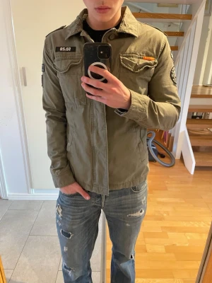 Olivgrön field jacket från Superdry - Säljer en olivgrön field jacket  från Superdry med coola patchar och broderade detaljer på ärmar och bröst. Två bröstfickor med lock och knappar, samt klassisk krage. Tillverkad i bomull för en skön och avslappnad look. (Modell: 177cm)