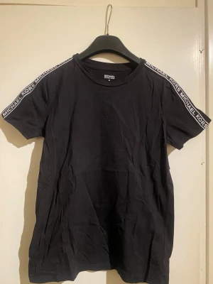 Svart Michael Kors t-shirt - Svart t-shirt från Michael Kors med korta ärmar och rund hals. Snygga logoband med texten 'MICHAEL KORS' längs axlarna ger en sportig vibe. Tillverkad i mjuk bomull för skön känsla och enkel passform.
