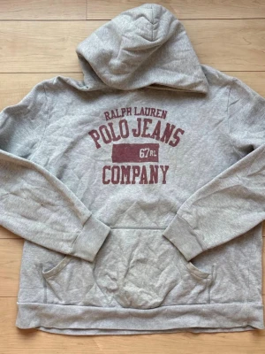 Grå hoodie från Polo Jeans Company - Snygg grå hoodie från Ralph Lauren Polo Jeans Company med stor magficka och tryck i vinrött framtill. Klassisk huva och ribbade muddar vid ärmslut och nederkant. Perfekt för en avslappnad och sportig stil.
