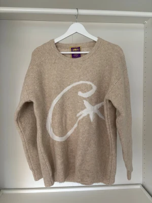 Beige stickad tröja från cortez - Stickad beige tröja från cortez med en stor vit grafisk detalj i form av ett C och en stjärna på framsidan. Tröjan har rund halsringning, långa ärmar och ribbade muddar vid ärmslut och nederkant. Perfekt för dig som gillar en avslappnad och trendig stil.