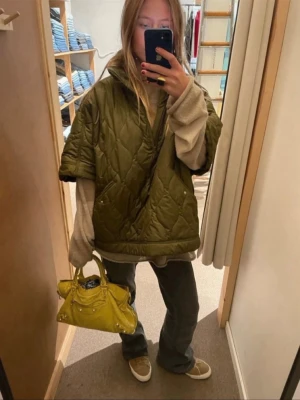 Olivgrön quiltad pufferjacka - Quiltad pufferjacka i olivgrönt med halv dragkedja och hög krage. Jackan har en loose passform och korta ärmar, perfekt för lager på lager. Tillverkad i polyester och har fickor framtill. Snygg och trendig jacka för dig som gillar streetstyle.