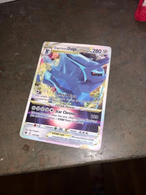 Origin Forme Dialga VSTAR Pokémonkort - Säljer ett Origin Forme Dialga VSTAR Pokémonkort med holografisk yta. Kortet har färger i blått, gult och silver och visar Dialga i sin VSTAR-form. Perfekt för samlare eller Pokémon TCG-spelare som vill ha en kraftfull och snygg kort i sin samling. Kortet är fejk