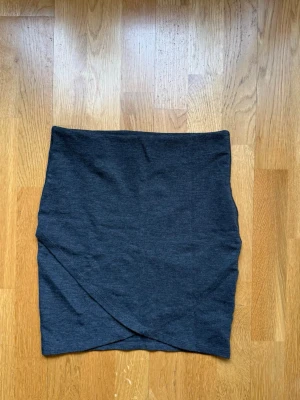 Mörkgrå omlottkjol i bomull - Mörkgrå kortkjol med omlottdetalj framtill. Kjolen är enkel och stilren, gjord i mjukt bomullsmaterial som känns bekvämt mot huden. Perfekt att matcha med en crop top eller hoodie för en avslappnad look.