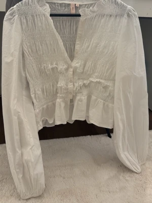 Vit smockad blus från Nelly - Söt vit blus från Nelly med smockdetaljer över bröstet och ryggen, volangkant nedtill och knappar framtill. Blusen har långa puffiga ärmar med resår vid ärmslut och en lätt rynkad krage. Perfekt för en trendig och romantisk look.