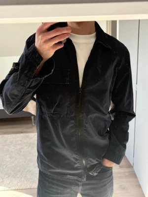 Marinblå overshirt från Stone Island - Stilren marinblå overshirt från Stone Island med dragkedja framtill och klassisk krage. Bröstficka på vänster sida och ikonisk logotyp-patch på vänster ärm. Tillverkad i bomull med en lätt tvättad finish som ger en avslappnad look.