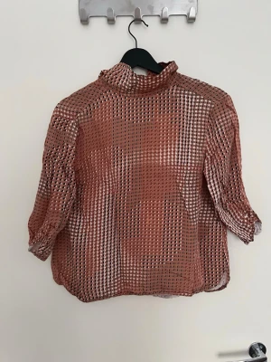 Rostfärgad transparent blus med mönster - Trendig rostfärgad blus i transparent material med rutigt mönster. Blusen har hög krage och trekvartsärmar. Perfekt för dig som gillar en luftig och unik stil. Materialet är lätt och ger en cool look.