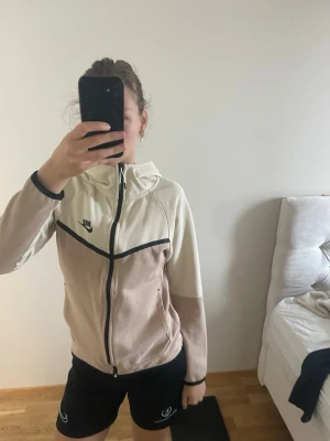 Nike beige och vit zip hoodie - Snygg zip hoodie från Nike i beige och vitt med svarta detaljer. Jackan har dragkedja framtill, huva och två sidofickor. Perfekt för träning eller chill. Nike-logga på bröstet och svart kantband ger en sportig look.