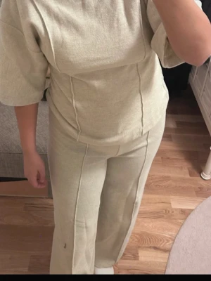 Beige loungewear set med vida ben - Mysigt loungewear set i beige med kortärmad topp och matchande byxor med vida ben. Setet har en avslappnad passform och är gjort i mjukt  material som passar perfekt för hemmakvällar såsom utekvällar. Snygga sömdetaljer framtill ger en modern touch.