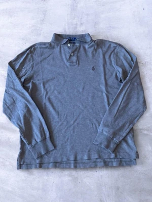 Brun långärmad piké från Polo Ralph Lauren - Klassisk brun långärmad piké från Polo Ralph Lauren med broderad logga på bröstet. Skjortan har knappar vid halsen och är tillverkad i mjuk bomull. Perfekt för en clean och avslappnad stil.