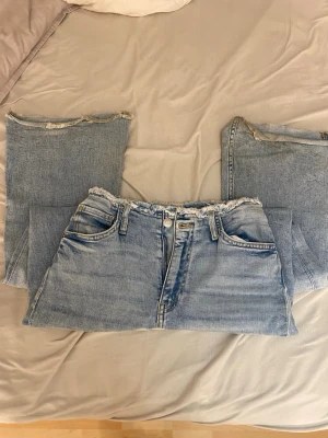 Ljusblåa jeans med råa kanter - Säljer ett par ljusblå bootcut jeans i klassisk femficksmodell med råa, avklippta kanter i midja och benslut. Jeansen har en avslappnad passform och är tillverkade i denim. Perfekta för en trendig och avslappnad stil. Storlek 158 passar för xxs/xs och även S tror jag