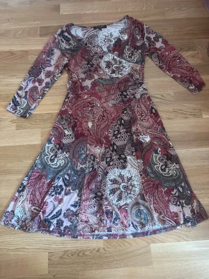 Mönstrad långklänning i paisley - Säljer en långklänning med paisley- och blommönster i vinröda, beige, svarta och blå toner. Klänningen har lång ärm, rund halsringning och är tillverkad i ett mjukt, stretchigt material som känns bekvämt mot huden. Perfekt för dig som gillar bohemiska vibes.