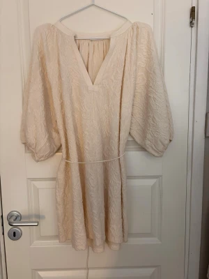Ljus kortklänning med v-ringning - Säljer en ljus kortklänning med v-ringning och vida trekvartsärmar. Klänningen har ett subtilt strukturmönster och ett smalt knytband i midjan för en snygg siluett. Perfekt för sommarens alla tillfällen!