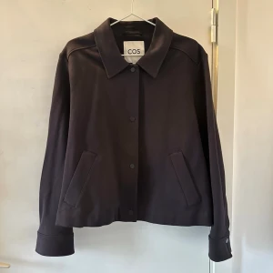 Mörkblå jacka med knappar från COS - Stilren mörkblå jacka från COS med krage och raka fickor framtill. Jackan har knappar längs hela framsidan. Perfekt om man tycker om oversize. Passar för unisex. Mycket bra skick💛
