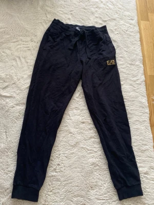 Svarta EA7 mjukisbyxor från Armani - Säljer ett par svarta mjukisbyxor från EA7 Emporio Armani med gul logga på vänster ben. Byxorna har dragsko i midjan och muddar vid bensluten. Perfekta för chill eller träning, riktigt sköna och stilrena. Den har ett litet hål vid knän 
