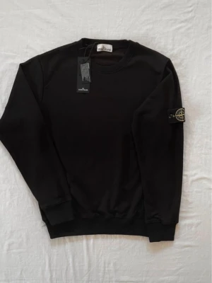 Svart sweatshirt från Stone Island - Svart sweatshirt från Stone Island med klassisk rund hals och ikonisk patch på vänster ärm. Tröjan har ribbade muddar vid ärmslut och nederkant. Perfekt för en clean och stilren look.